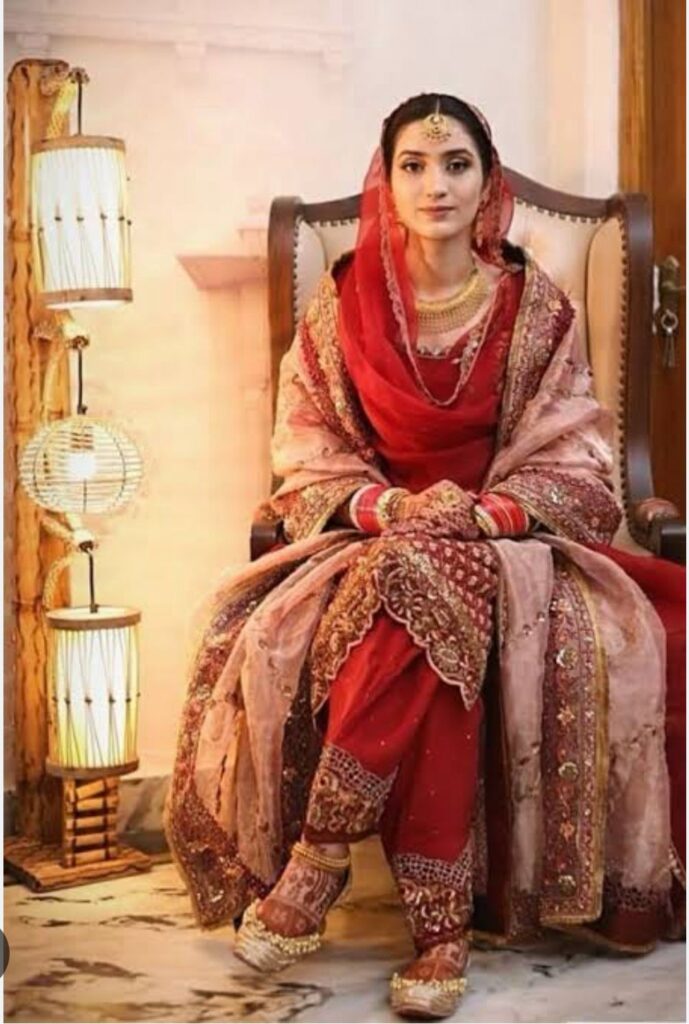 A royal Indian bride in a traditional red and gold bridal suit, seated elegantly with embellished Punjabi juttis, adorned with bridal jewelry and chooda, surrounded by warm ambient lighting.